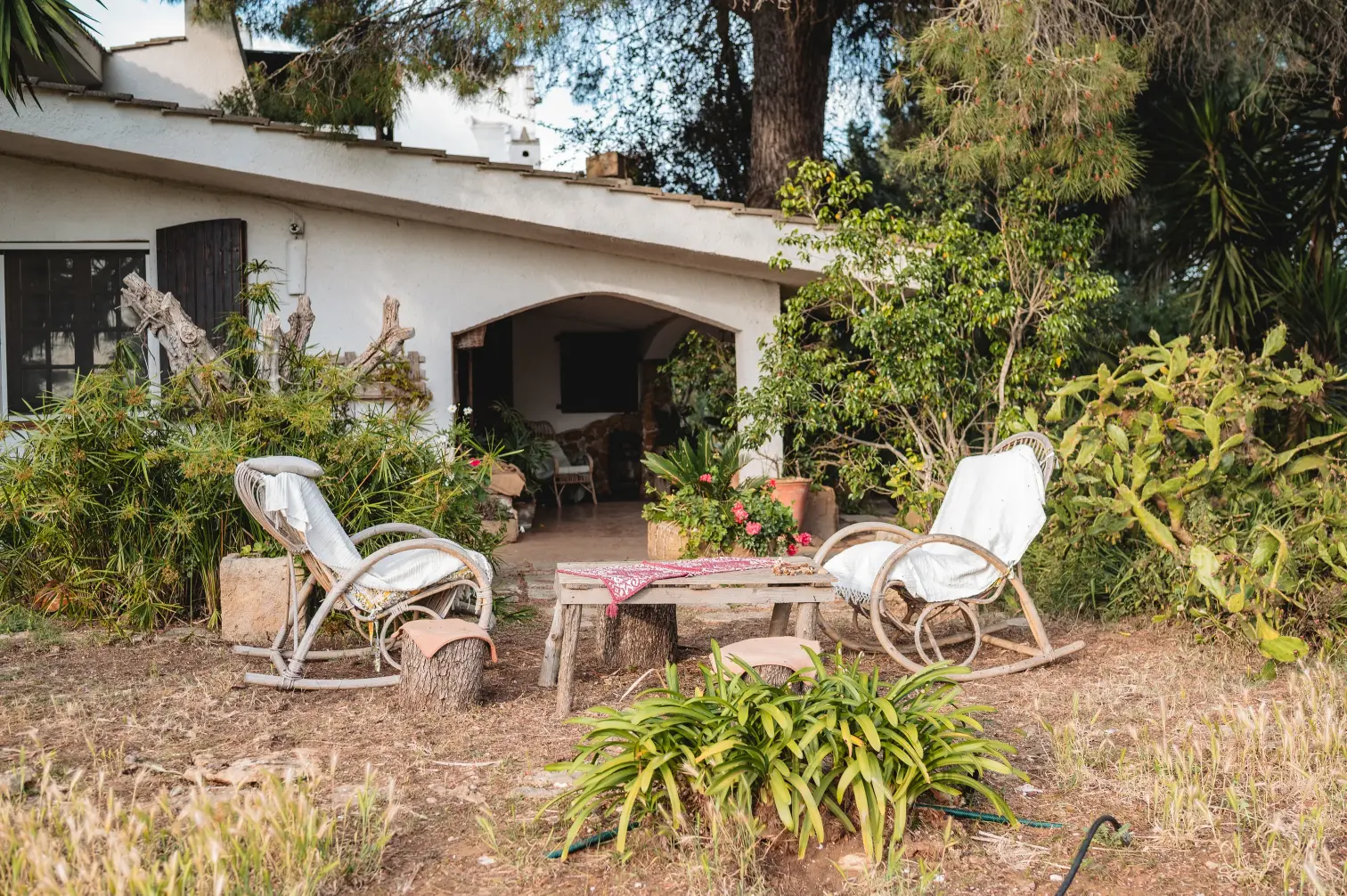 Giardino di una masseria con due sedie a dondolo in vimini, tavolino rustico e piante grasse sotto il porticato bianco