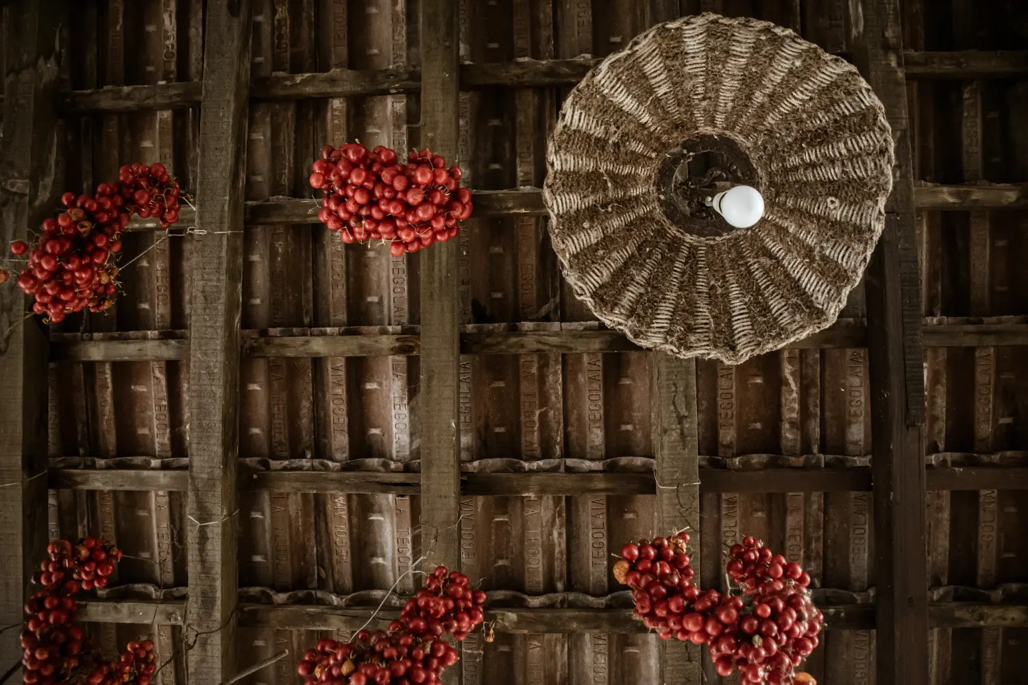 Dettaglio di un soffitto in tegole e travi di legno con grappoli di pomodori appesi a seccare e lampadario in paglia intrecciata