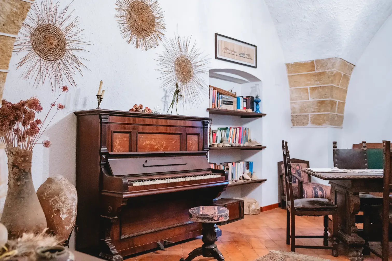 Interno rustico d'epoca con pianoforte verticale antico, sgabello, vasi in terracotta, librerie a muro e area pranzo in legno scolpito